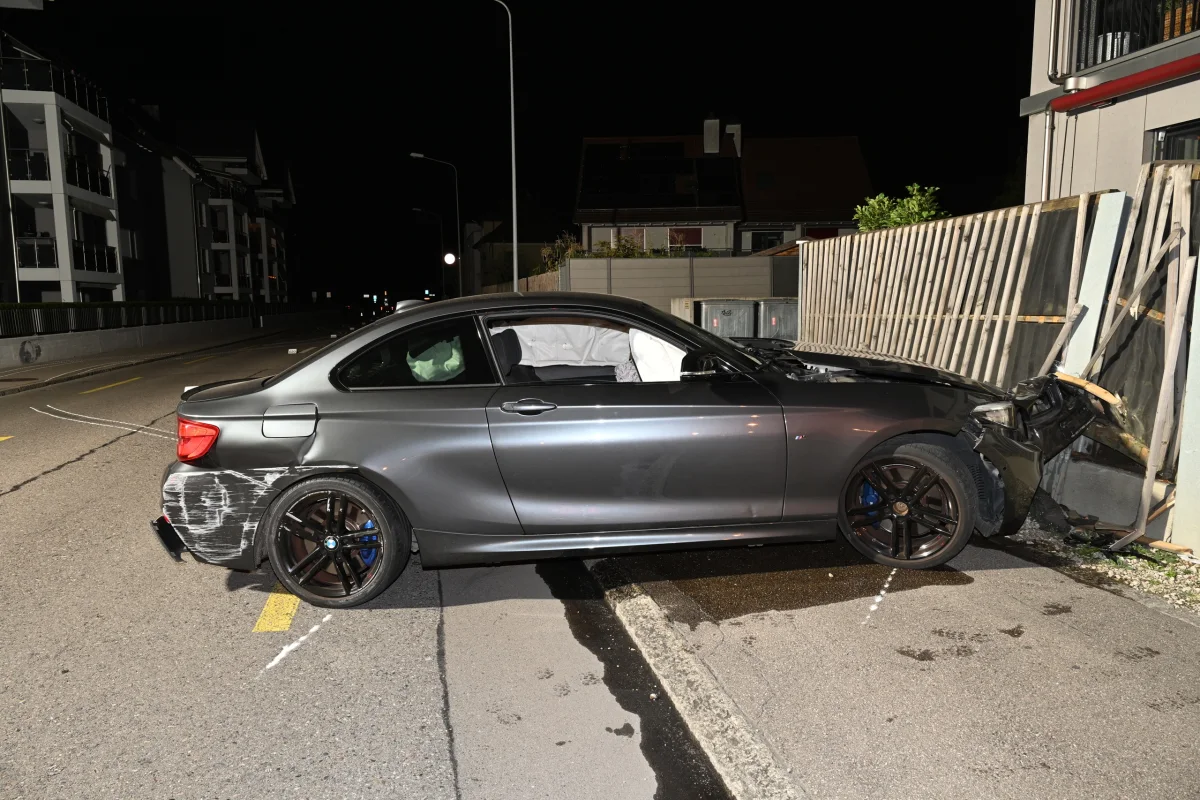 Ein silberner BMW steht mit deutlich sichtbarem Frontschaden vor einem beschädigten Zaun. Die Straße ist nachts leer.
