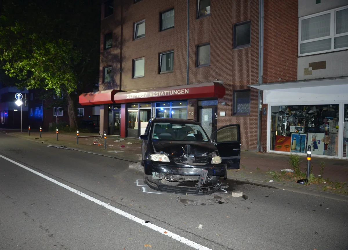 Ein schwarzes Auto mit Frontschaden und offener Tür steht vor einem Bestattungsinstitut auf einer Straße bei Nacht.