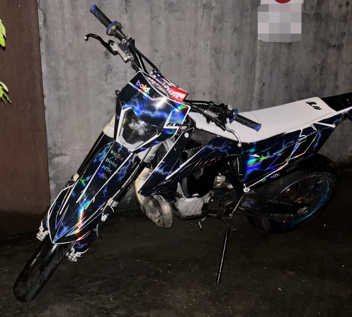 Ein dekoratives Motorrad mit buntem Design lehnt an einer grauen Wand, aufgenommen in einer dunklen Umgebung.