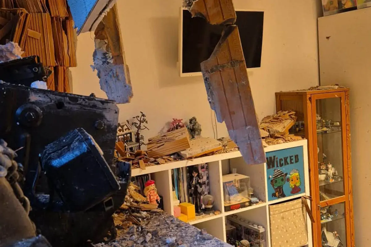 Ein Wohnzimmer mit schweren Schäden: Herabgefallene Trümmer und zerstörte Wände sind sichtbar. Regale voll mit persönlichen Gegenständen und Dekorationen sind betroffen.