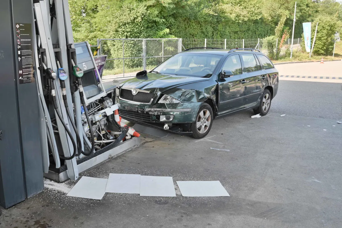 Ein grünes Auto ist an einer Tankstelle mit einer Zapfsäule kollidiert, was zu sichtbaren Schäden geführt hat. Kraftstoffleitungen und elektronische Bauteile der Säule sind durcheinandergebracht.