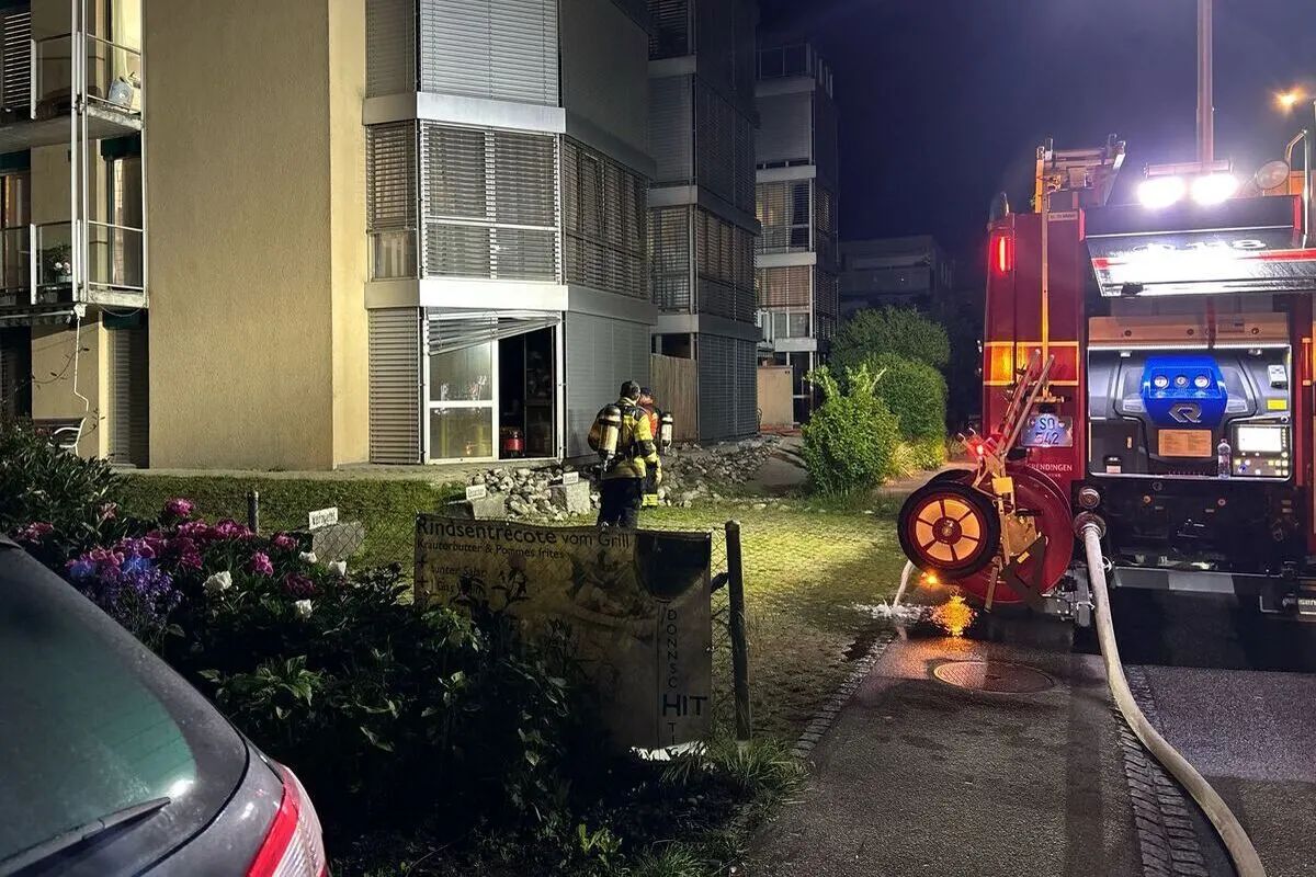 Ein beleuchtetes Feuerwehrfahrzeug steht mit Schlauch ausgelegt auf einer Straße. Zwei Feuerwehrleute stehen vor einem Wohnhaus.