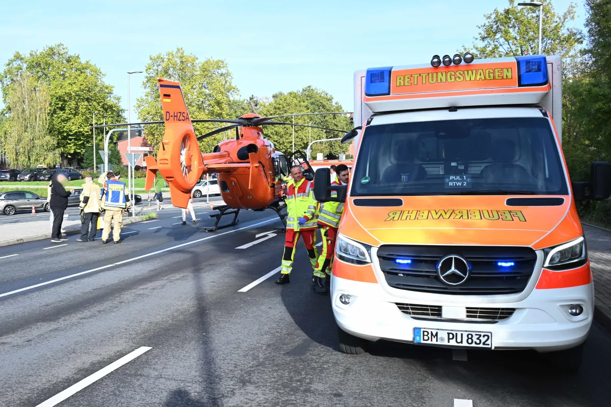 Ein oranger Rettungshubschrauber steht auf einer Straße neben einem Rettungswagen. Einsatzkräfte in Warnkleidung sind vor Ort.