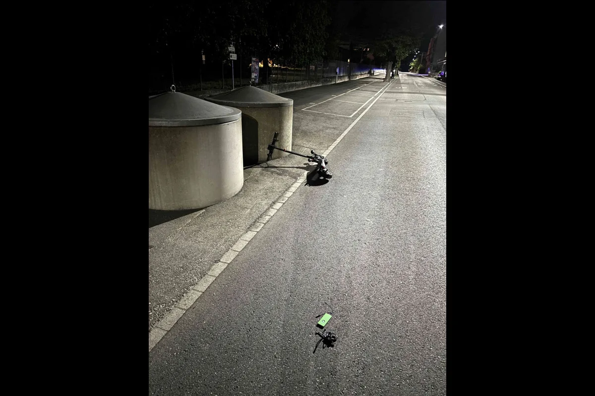 Zwei Elektro-Scooter liegen auf einem Gehweg neben zwei großen, runden Müllcontainern. Es ist nachts und die Straße ist leer.