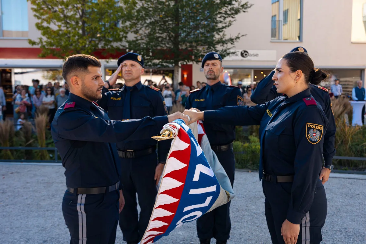 Mehrere Polizeibeamte in Uniform stehen zusammen, halten eine Fahne und nehmen an einer Zeremonie teil. Im Hintergrund stehen Zuschauer.