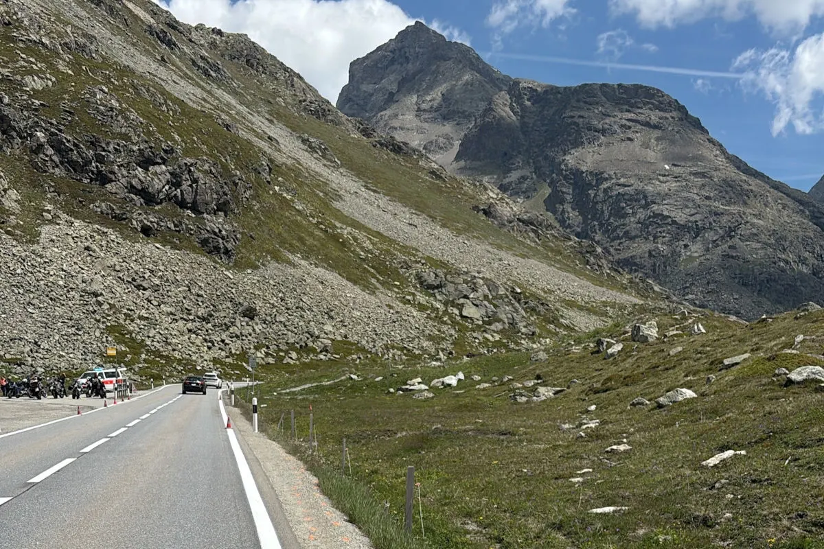 Eine Bergstraße führt an einem Berghang vorbei, wo mehrere Motorräder und ein Auto angehalten haben. Der Himmel ist klar und sonnig.