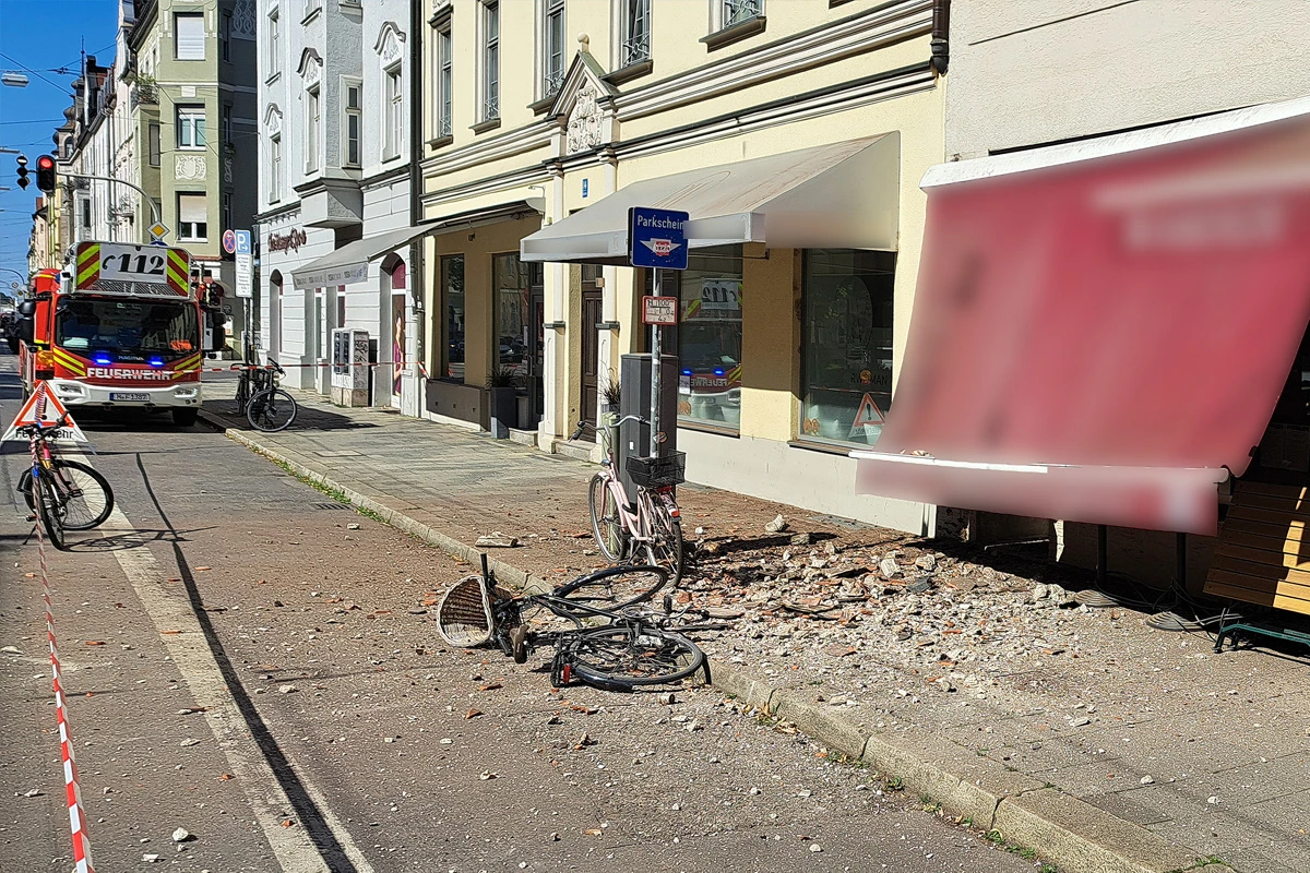 Ein zerstörtes Fahrrad liegt neben Trümmern auf dem Gehweg vor einem beschädigten Gebäude. Ein Einsatzfahrzeug der Feuerwehr ist im Hintergrund zu sehen.