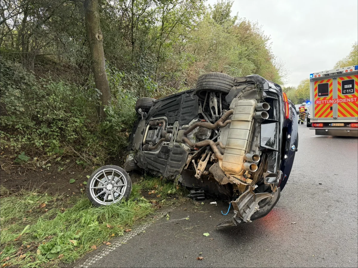 Ein blauer BMW liegt umgestürzt auf der Seite neben einer Straße. Ein Rettungswagen steht in der Nähe, umgeben von Grünflächen.