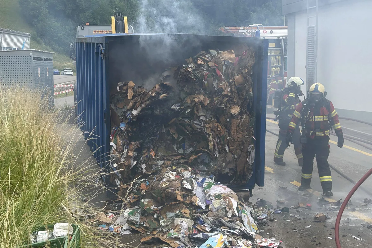 Ein Container voller brennender, rauchender Papiere wird von Feuerwehrleuten mit Atemschutzgeräten gelöscht. Zwei Feuerwehrleute stehen rechts im Bild neben dem Rauch.