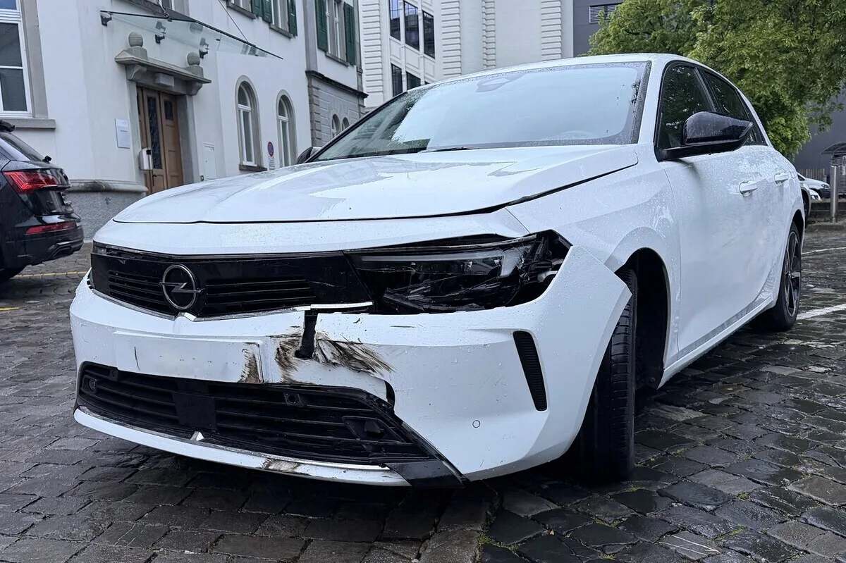 Ein weißer Opel mit deutlichen Schäden an der Vorderseite, insbesondere am Kühlergrill und an der Stoßstange, steht auf einer gepflasterten Straße vor einem älteren Gebäude.