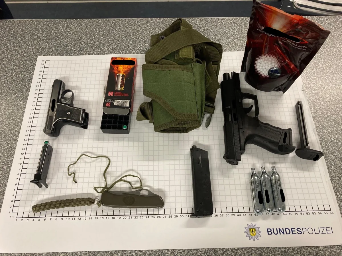 Auf einem Tisch liegen zwei Schreckschusspistolen, Verpackungen für Munition, CO2-Patronen, ein Messer und anderer Zubehör auf einem Diagramm der Bundespolizei.