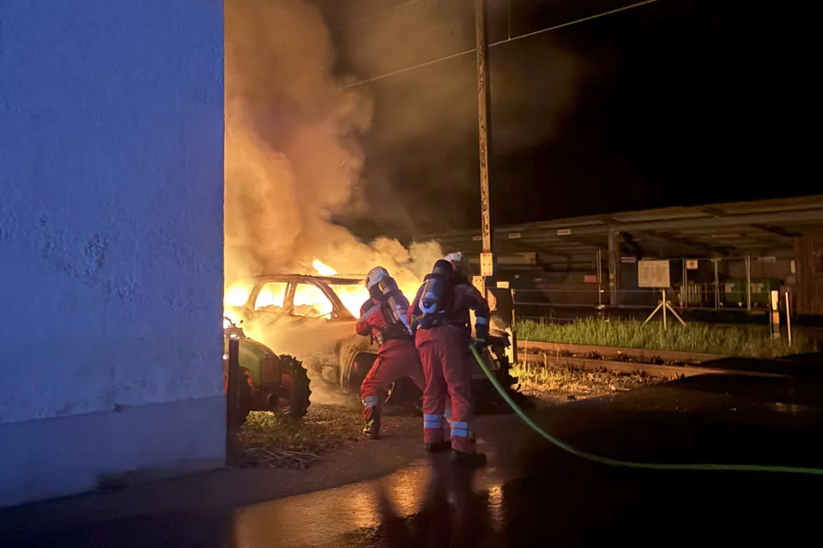Zwei Feuerwehrleute in Einsatzkleidung bekämpfen einen nächtlichen Fahrzeugbrand. Flammen schlagen aus dem Auto, während sie Wasser aufspritzen.