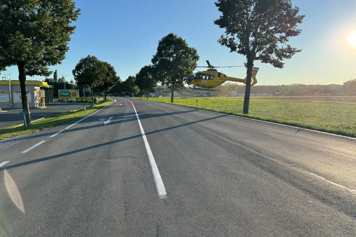 Ein gelber ADAC-Rettungshubschrauber schwebt über einer Wiese, während er neben einer ruhigen Landstraße landet. Die Sonne scheint über die Landschaft, und die Straße ist von Bäumen gesäumt.