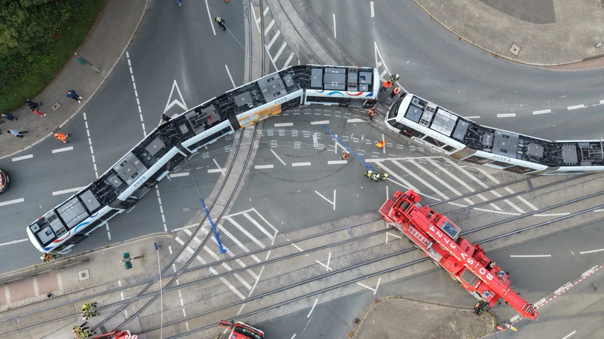 Luftaufnahme einer entgleisten Straßenbahn an einer Kreuzung, umgeben von Einsatzkräften und einem roten Abschleppfahrzeug. Mehrere Personen tragen orangefarbene Warnwesten.