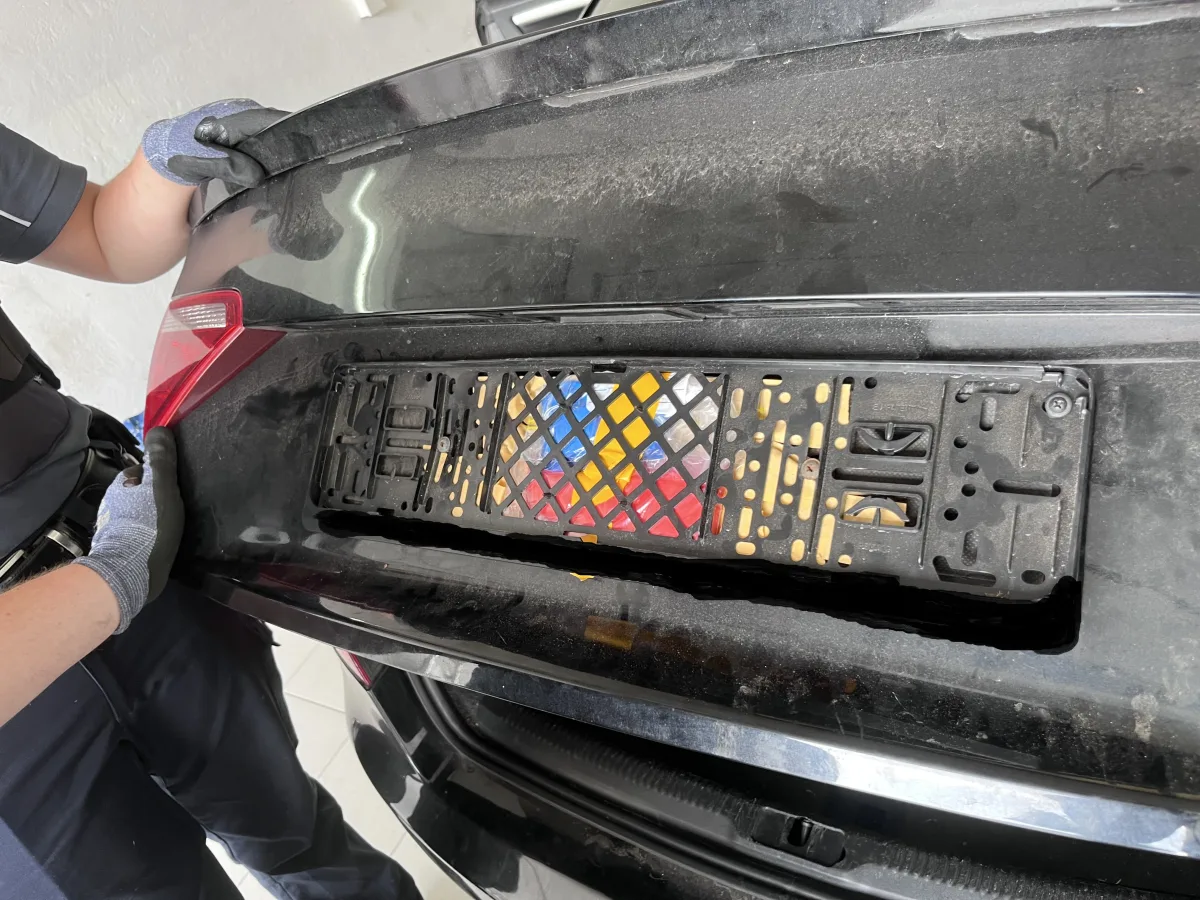 Ein geheimfach hinter einer farbigen Kennzeichenhalterung auf einem schwarzen Auto wird von einer Person geöffnet.