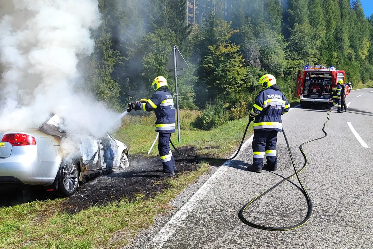 Zwei Feuerwehrmänner in voller Montur löschen ein stark qualmendes Auto am Straßenrand. Im Hintergrund steht ein Feuerwehrfahrzeug.