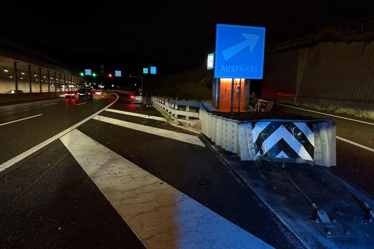 Ein Auto ist bei Nacht an einer Autobahnausfahrt in die Leitplanke gefahren. Die Umgebung ist durch Verkehrsschilder und beleuchtete Ausfahrtszeichen gekennzeichnet.
