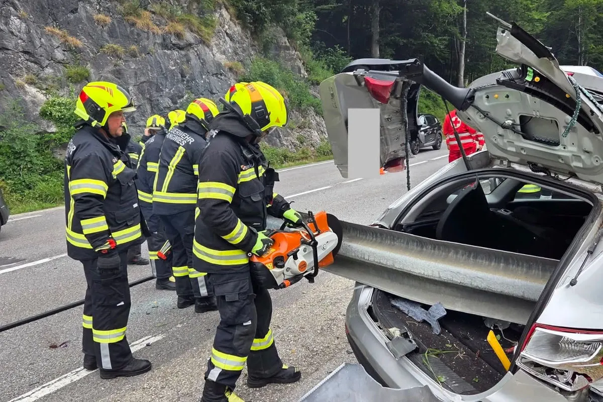 Feuerwehrmänner in Schutzkleidung arbeiten an einem Auto, dessen Heck in eine Leitplanke gerammt ist. Das Fahrzeug ist stark beschädigt.