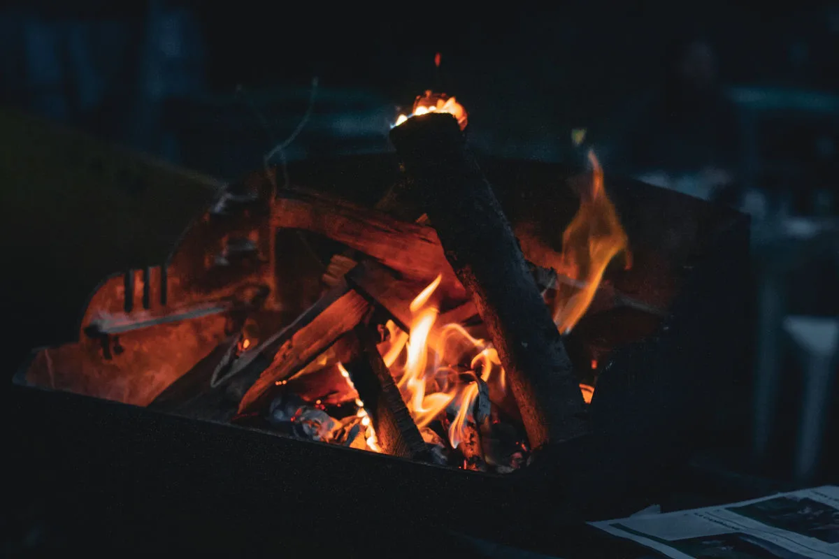 Ein Feuer brennt in einem Metalbehälter und Holzscheite befinden sich in den Flammen. Die Dunkelheit umgibt das Bild.