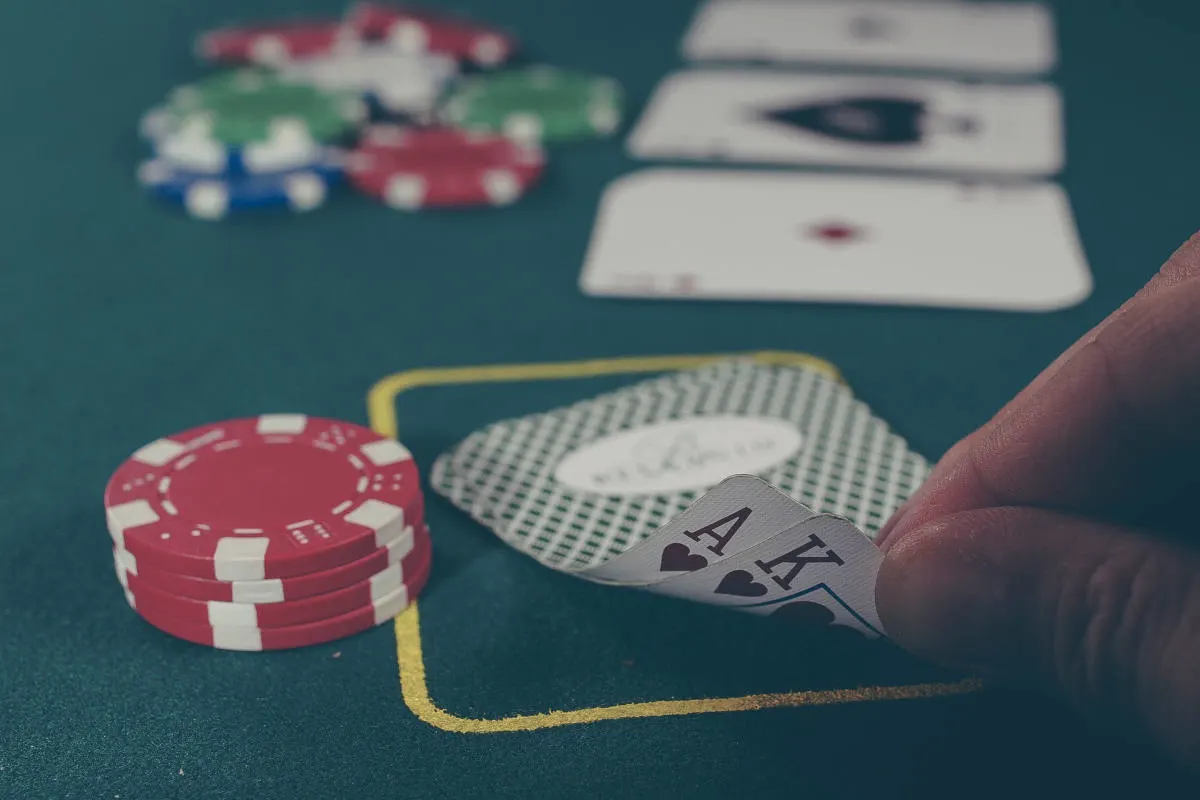 Auf einem Pokertisch liegen rote Pokerchips und eine Hand zeigt ein Ass und einen König in Herz. Im Hintergrund liegen Karten offen auf dem Tisch.