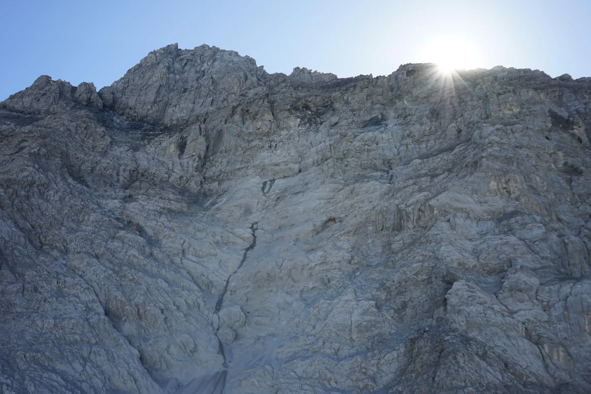 Ein felsiger Berggipfel, über dem die Sonne strahlt, erzeugt helle Lichtstrahlen. Der Himmel ist klar.