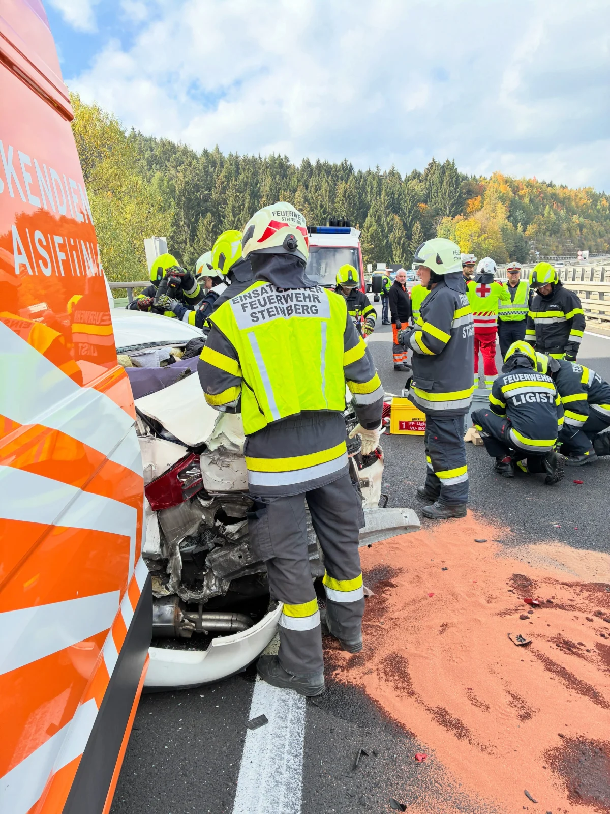 Mehrere Feuerwehrleute stehen um ein beschädigtes Auto auf einer Autobahn. Im Hintergrund sind Bäume zu sehen.