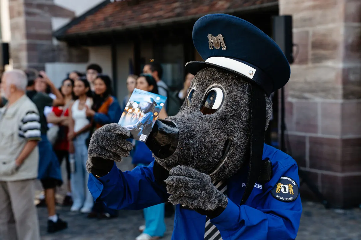 Ein Polizei-Maskottchen der Kantonspolizei Basel-Stadt hält ein Foto in den Händen, umgeben von einer Gruppe von Menschen im Freien.