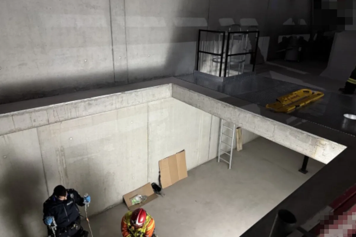 Zwei Rettungskräfte befinden sich in einem leeren Betonschacht mit einer Leiter und Rettungsausrüstung. Die Szene spielt in einem Innenraum.