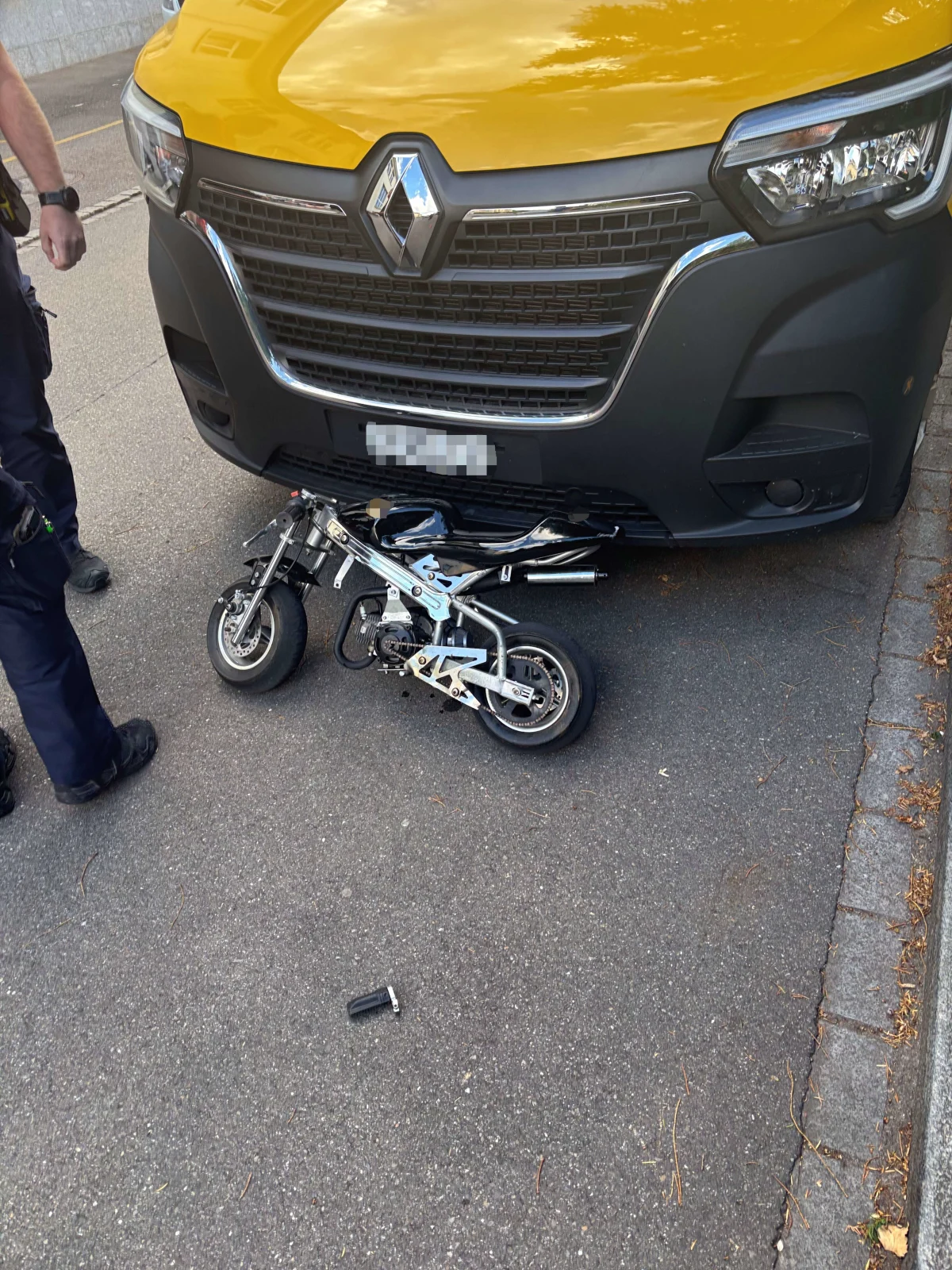 Ein gelber Renault-Van steht auf der Straße, unter dessen Front ein kleines Motorrad eingeklemmt ist. Ein Mann in dunkler Kleidung steht daneben.
