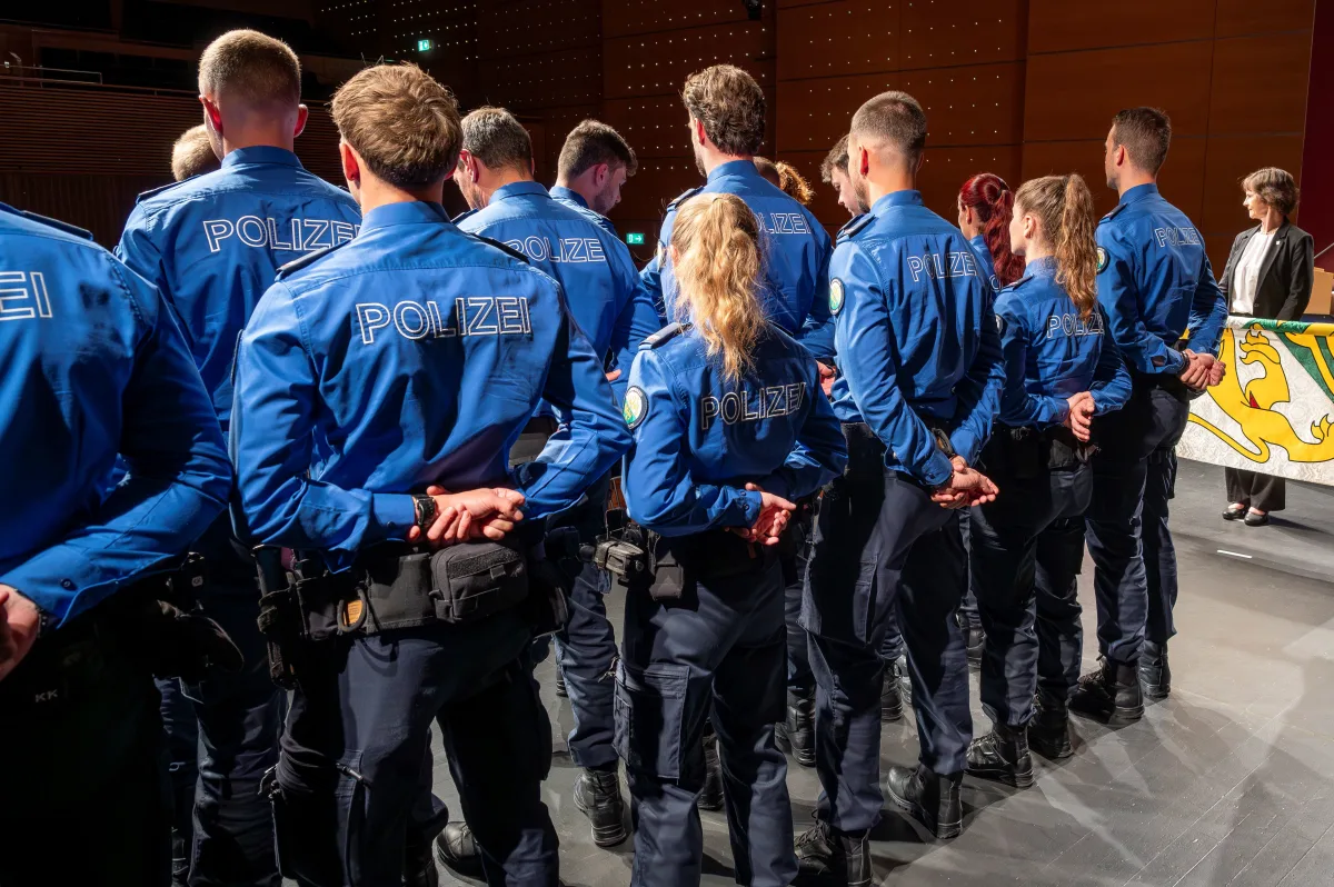 Mehrere Polizisten stehen in blauer Uniform mit dem Rücken zur Kamera. Eine Frau steht an einem Rednerpult im Hintergrund.