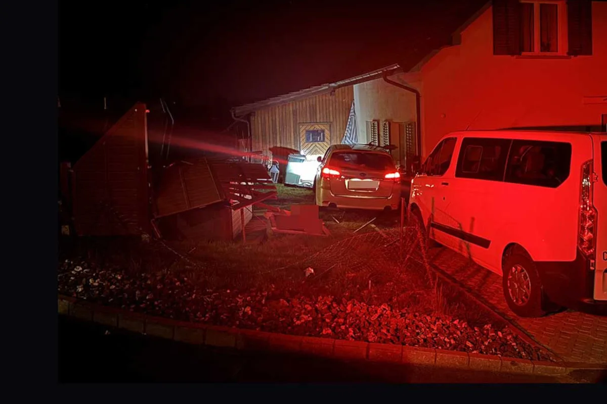 Bei Nacht sind zwei Autos vor einem Gebäude zu sehen, eines mit eingeschaltetem Rücklicht. Der Vorgarten und der Zaun sind beschädigt.