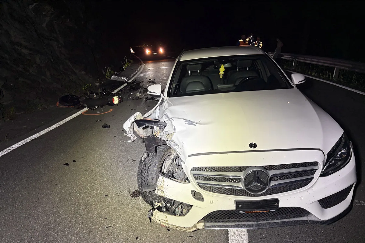 Ein weißer Mercedes mit schweren Beschädigungen an der Front steht auf einer dunklen Straße. Im Hintergrund sind Unfalltrümmer sichtbar.