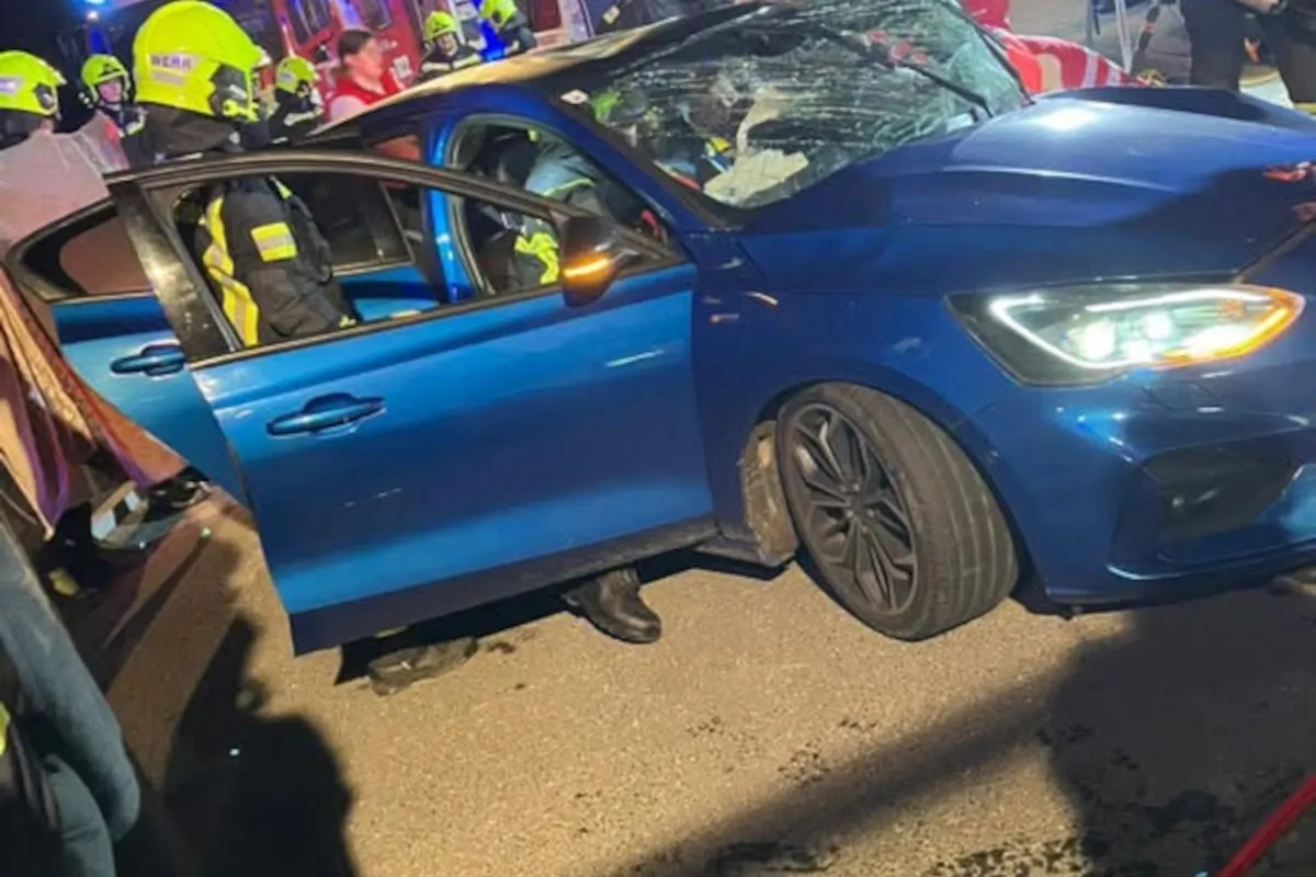 Ein blauer Pkw mit beschädigter Windschutzscheibe steht auf der Straße, umgeben von Feuerwehrleuten in gelben Helmen bei Nacht.