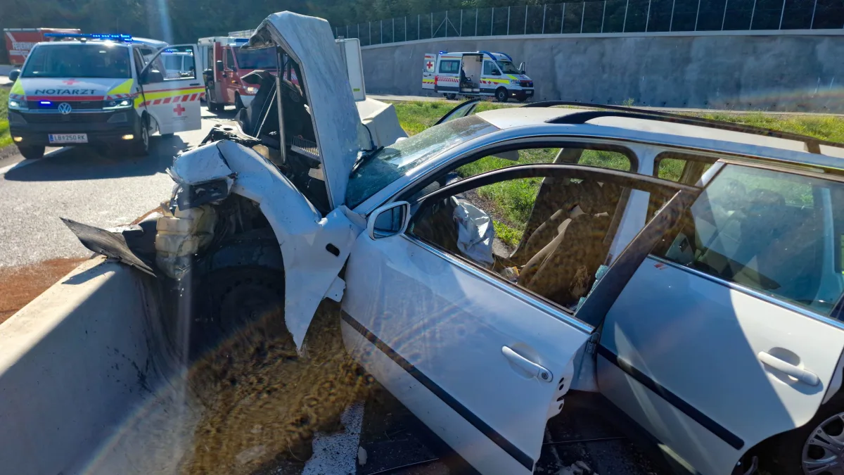 Ein stark beschädigtes, weißes Auto steht an einer Betonbarriere. Im Hintergrund sind Rettungsfahrzeuge sichtbar.