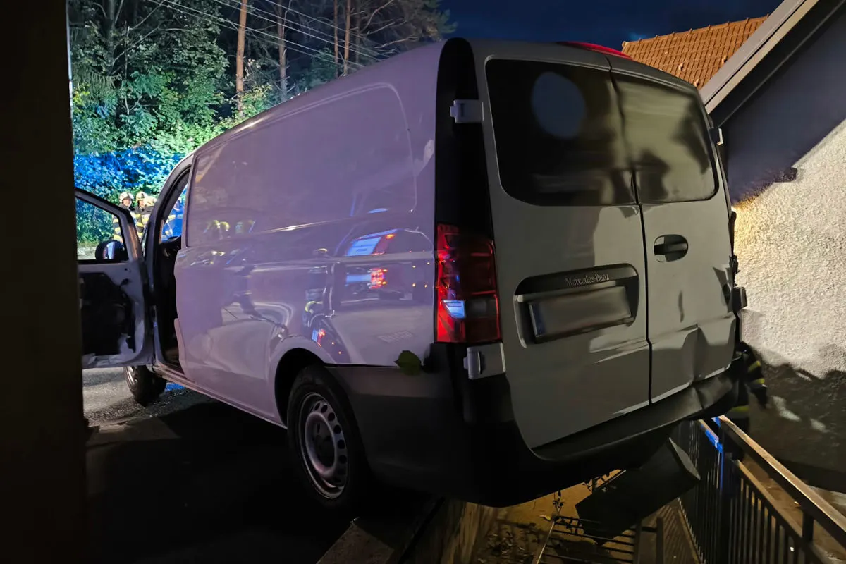 Ein weißer Mercedes-Transporter steht mit dem Heck über einem Geländer, als ob er hinunterstürzen könnte. Es ist Nacht, und im Hintergrund sind Bäume zu sehen.