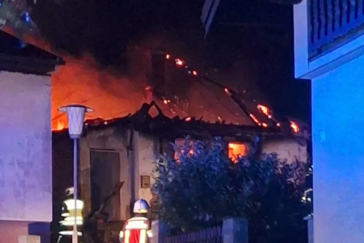 Ein Haus steht bei Nacht in Flammen, das Dach ist von Feuer und Rauch bedeckt. Zwei Einsatzkräfte in Schutzkleidung sind vor Ort.