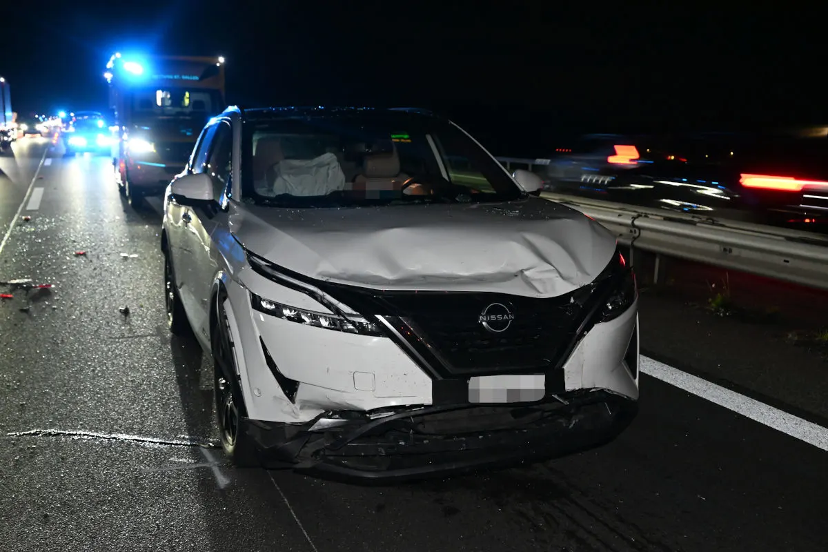 Ein weißer Nissan mit erheblichen Frontschäden steht bei Nacht auf einer Autobahn. Im Hintergrund sind Einsatzfahrzeuge zu erkennen.