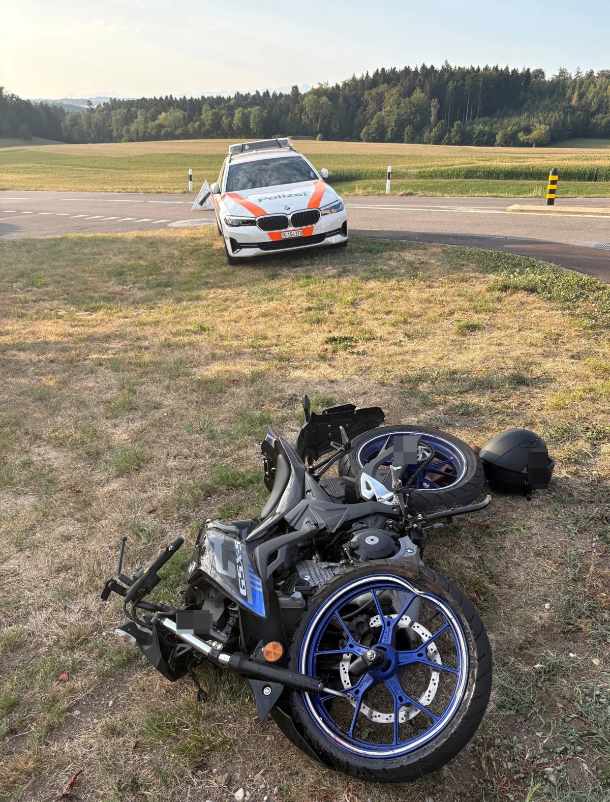 Auf einer Wiese liegt ein beschädigtes Motorrad. Im Hintergrund steht ein Polizeiauto nahe einer Landstraße vor einem Wald.