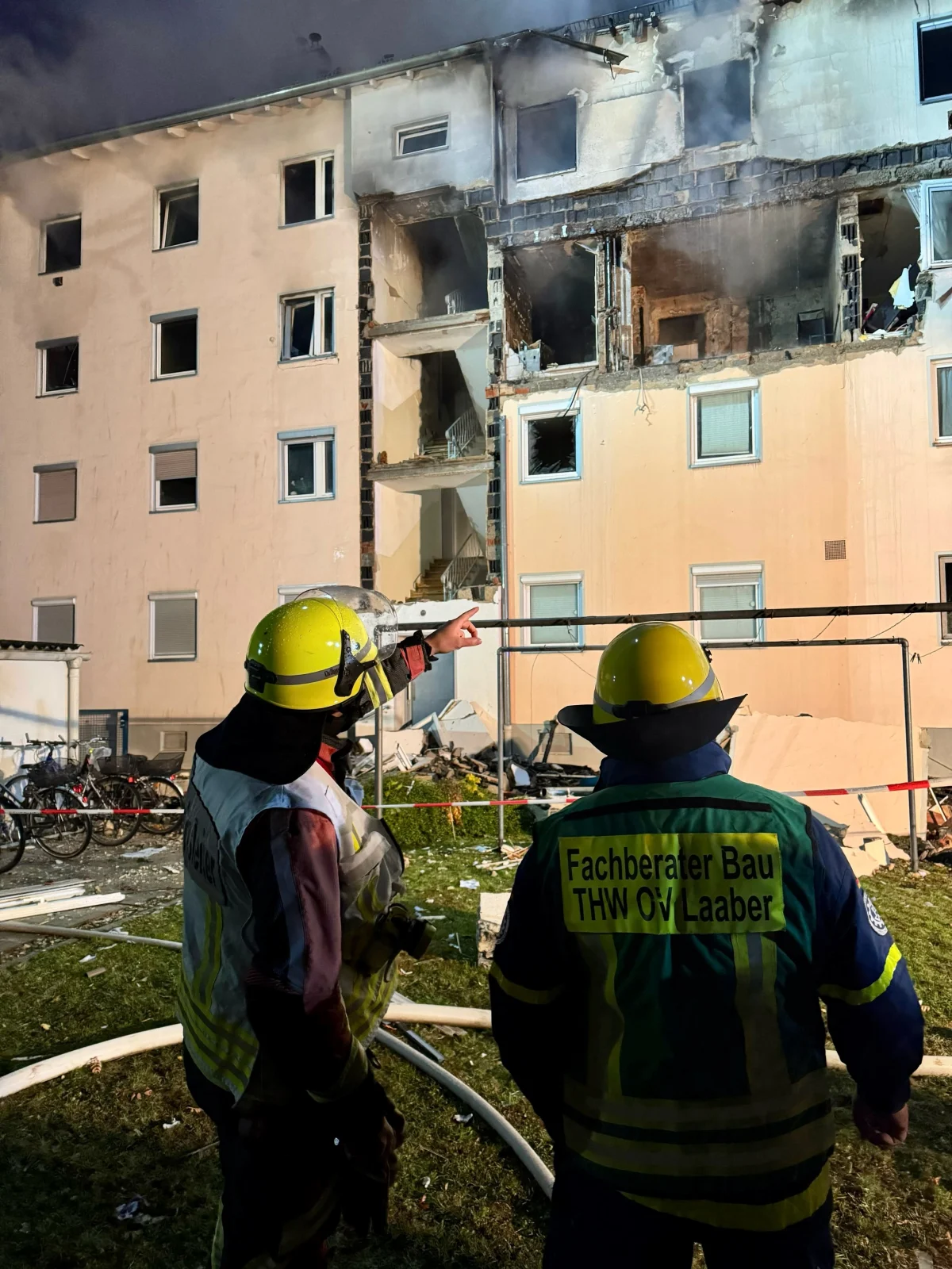 Zwei Feuerwehrleute stehen vor einem beschädigten Mehrfamilienhaus. Das Gebäude zeigt deutliche Brandspuren und ist teilweise zerstört.
