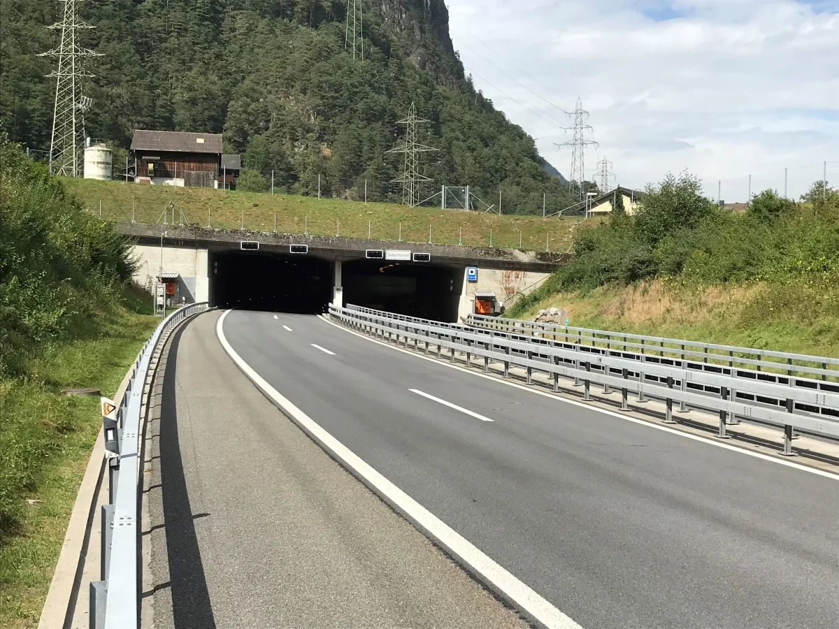 Ein leeres Autobahnstück führt in einen Tunnel, der von Bergen und Vegetation umrahmt ist. Stromleitungen ziehen sich über die Landschaft.