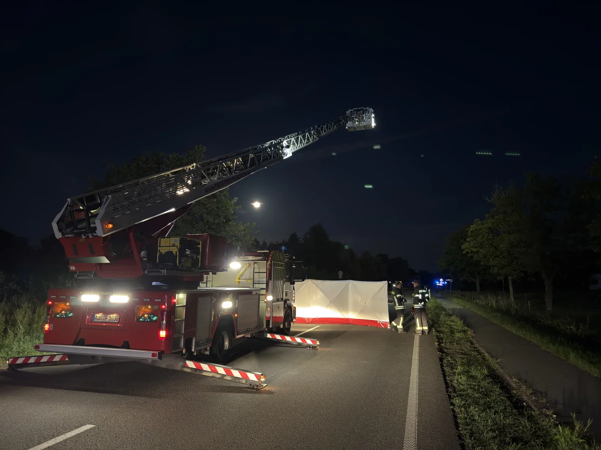 Ein rotes Feuerwehrfahrzeug steht nachts mit ausgefahrener Drehleiter auf einer Landstraße. Mehrere Feuerwehrleute sind in der Nähe einer weißen Abschirmung zu sehen.