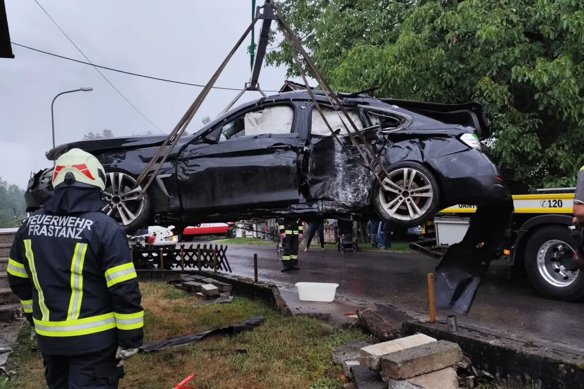 Ein schwarzes, beschädigtes Auto wird mit einem Kran angehoben. Im Vordergrund ist ein Feuerwehrmann zu sehen, der die Bergung überwacht. Die Szenerie spielt sich auf einer nassen Straße ab.