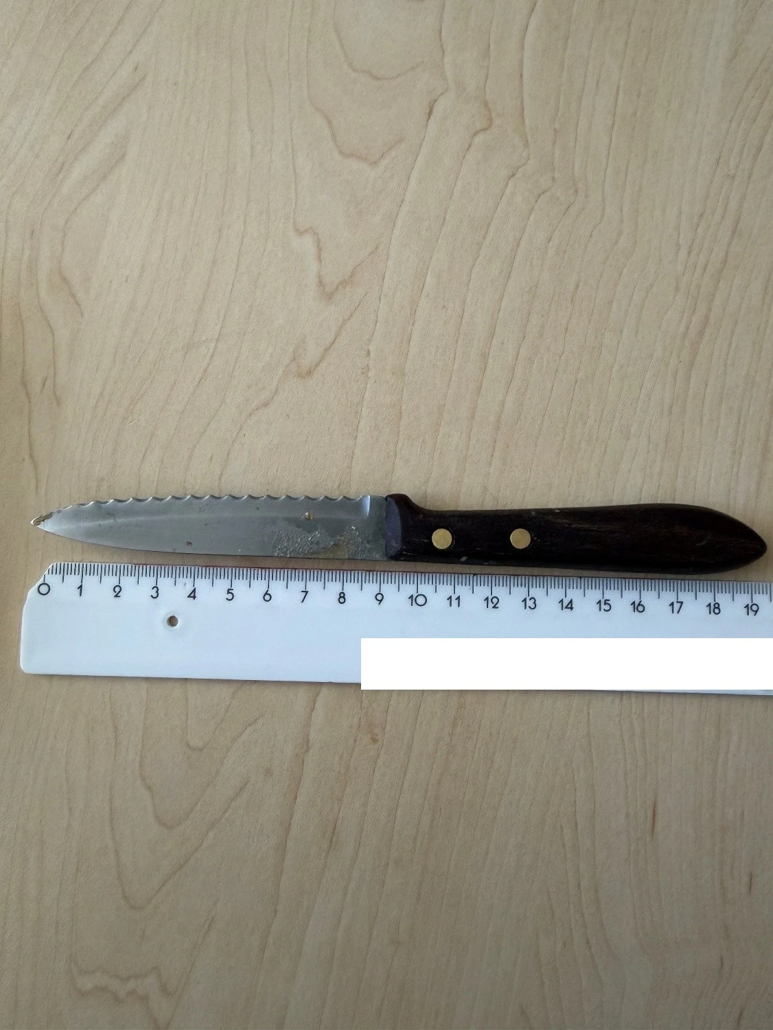 Ein Messer mit Wellenschliff liegt auf einem Holztisch neben einem Lineal, das die Größe anzeigt.