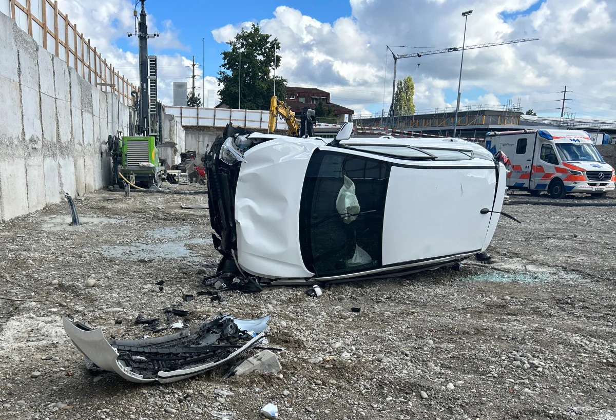 Ein weißes Auto liegt seitlich auf einer Baustelle. Ein Krankenwagen ist im Hintergrund zu sehen, bei bewölktem Himmel.