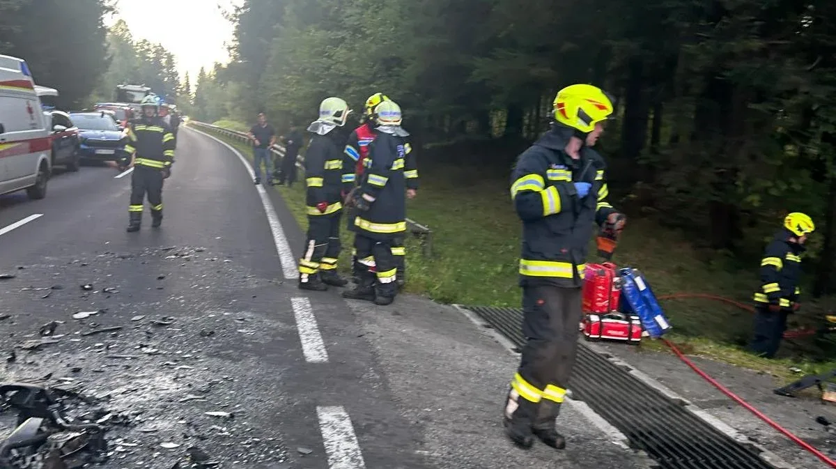 Mehrere Feuerwehrleute in Schutzkleidung stehen auf einer Landstraße nahe einer Unfallstelle. Trümmer sind auf der Fahrbahn sichtbar. Ein Rettungswagen und weitere Fahrzeuge stehen im Hintergrund.