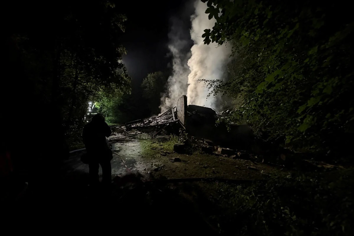 In einem bewaldeten Gebiet sind die Überreste einer Holzhütte zu sehen, aus denen Rauch aufsteigt. Die Szene ist in der Nacht aufgenommen.