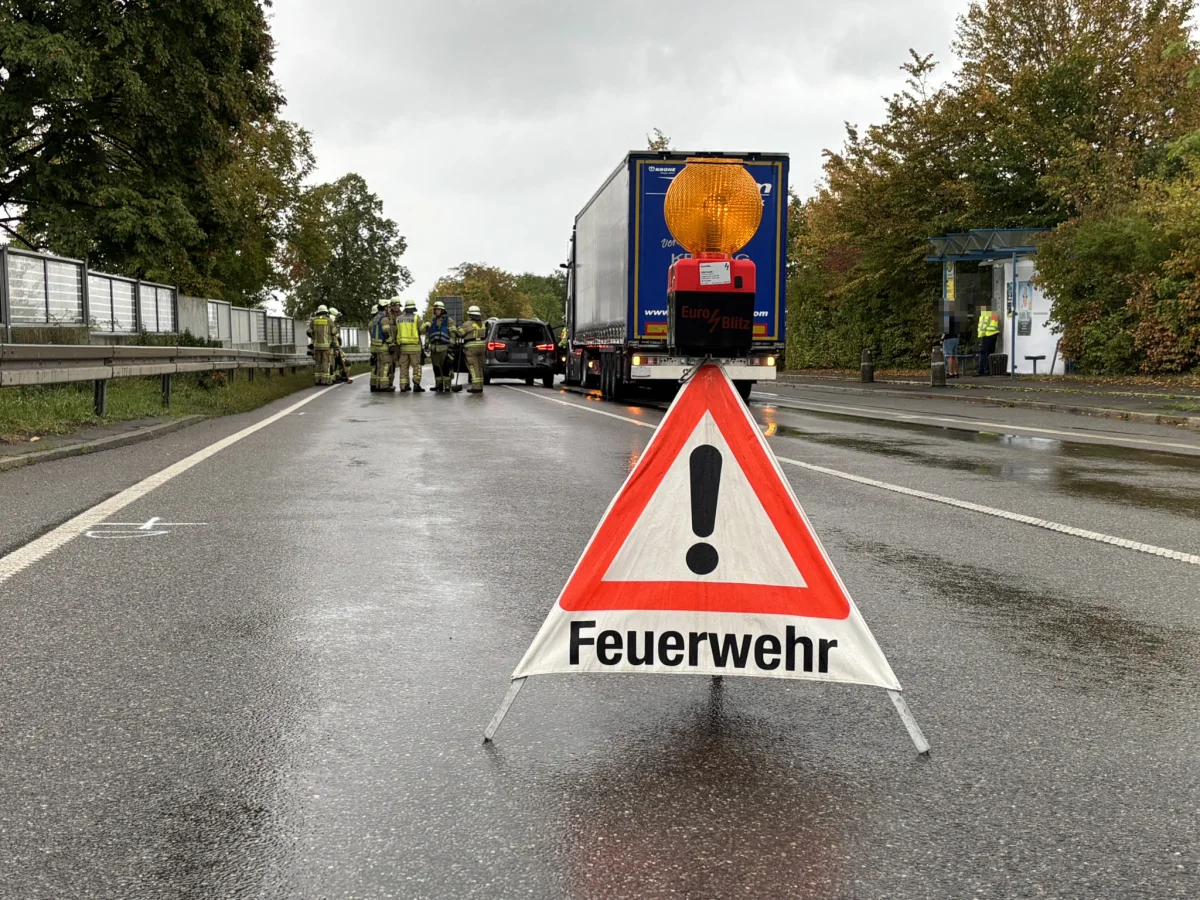 Ein Warnschild mit der Aufschrift 'Feuerwehr' steht auf einer regennassen Straße. Im Hintergrund sind Einsatzkräfte und ein Lkw zu sehen.