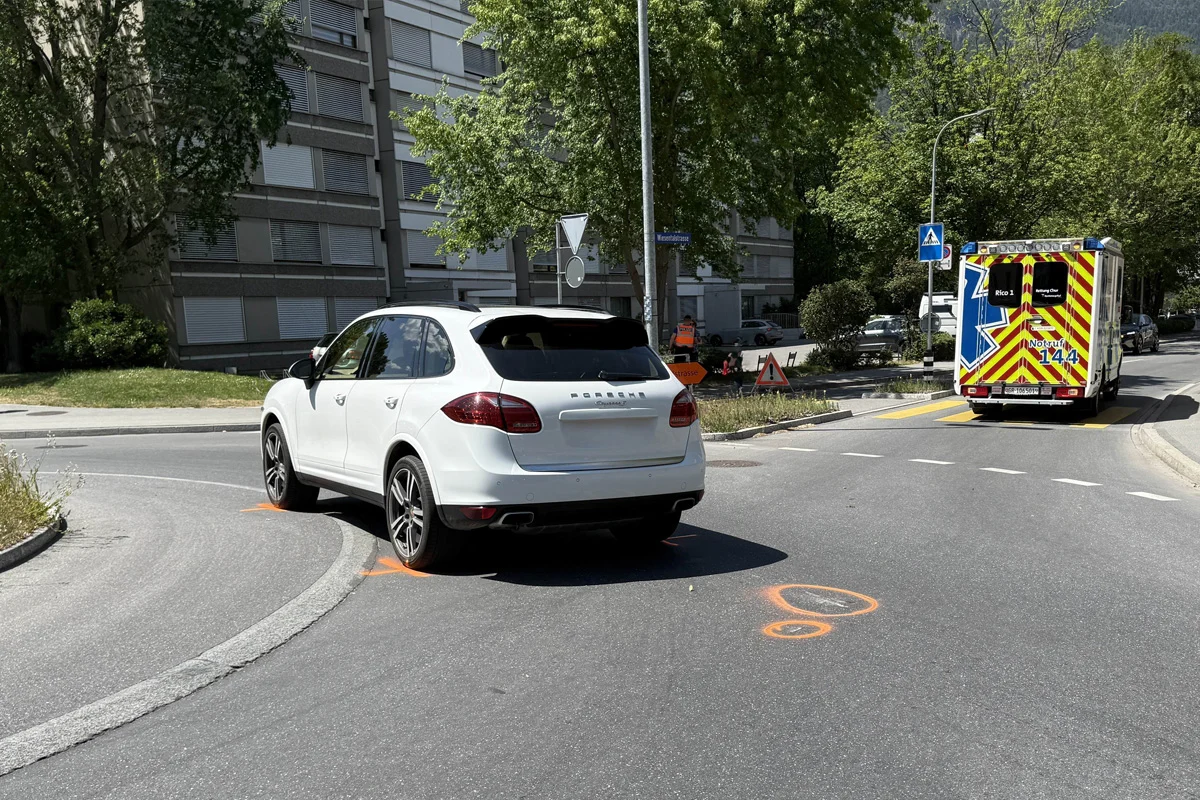 Ein weißes Auto steht in der Nähe eines Bordsteins, während ein Ambulanzfahrzeug mit auffälliger Warnmarkierung auf der Straße fährt. Wohngebäude und Bäume sind im Hintergrund sichtbar.