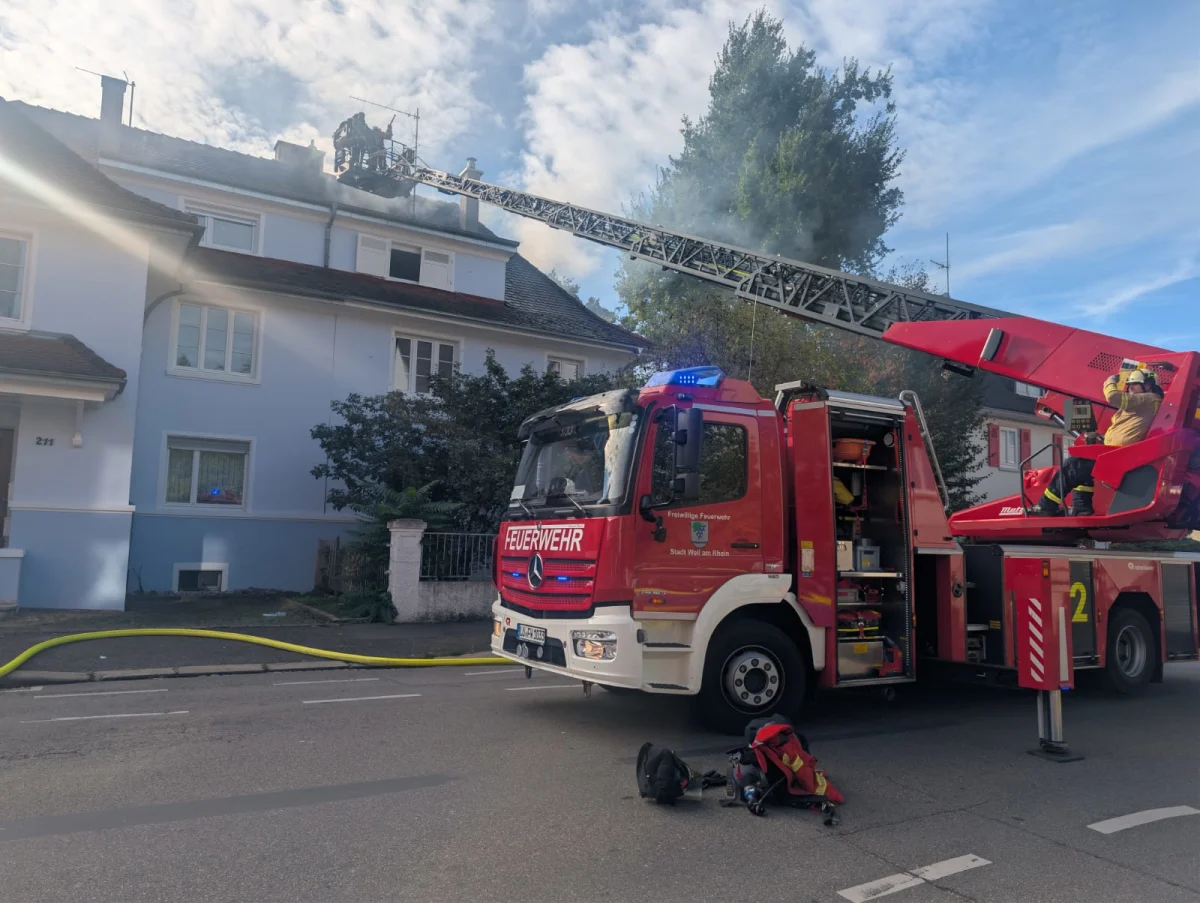 Ein rotes Feuerwehrfahrzeug steht vor einem Wohnhaus, während eine Leiter zum Dachstuhl ausgefahren ist. Feuerwehrleute bekämpfen den Brand von oben.