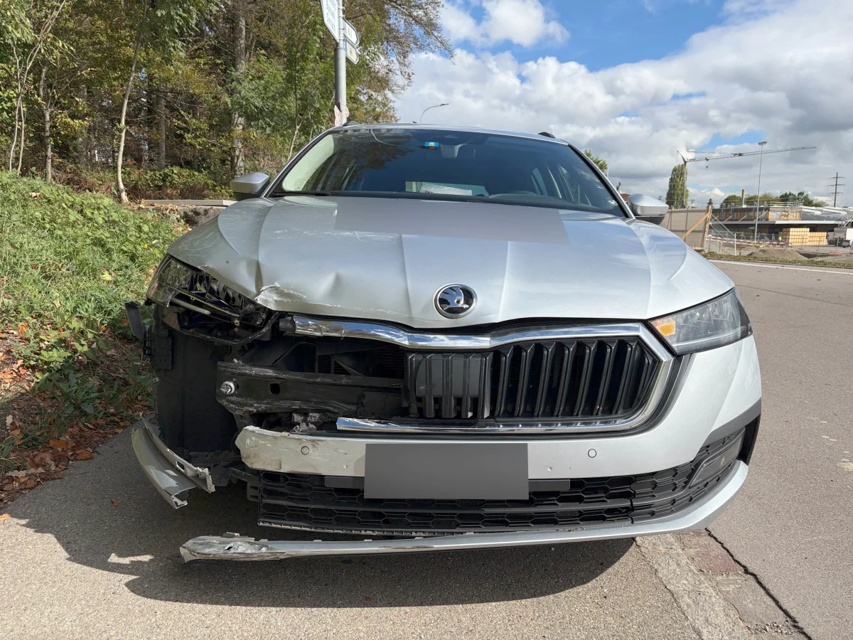 Ein silbernes Auto mit stark beschädigter Front steht am Straßenrand. Im Hintergrund sind Bäume und eine Baustelle sichtbar.